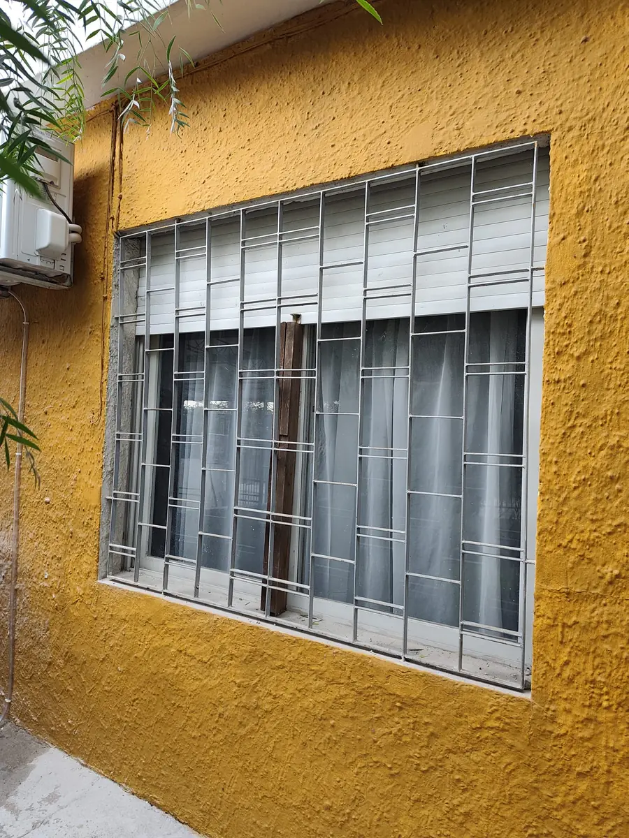 Cortina de enrollar blanca instalada en ventana con reja de hierro artesanal, pared color mostaza